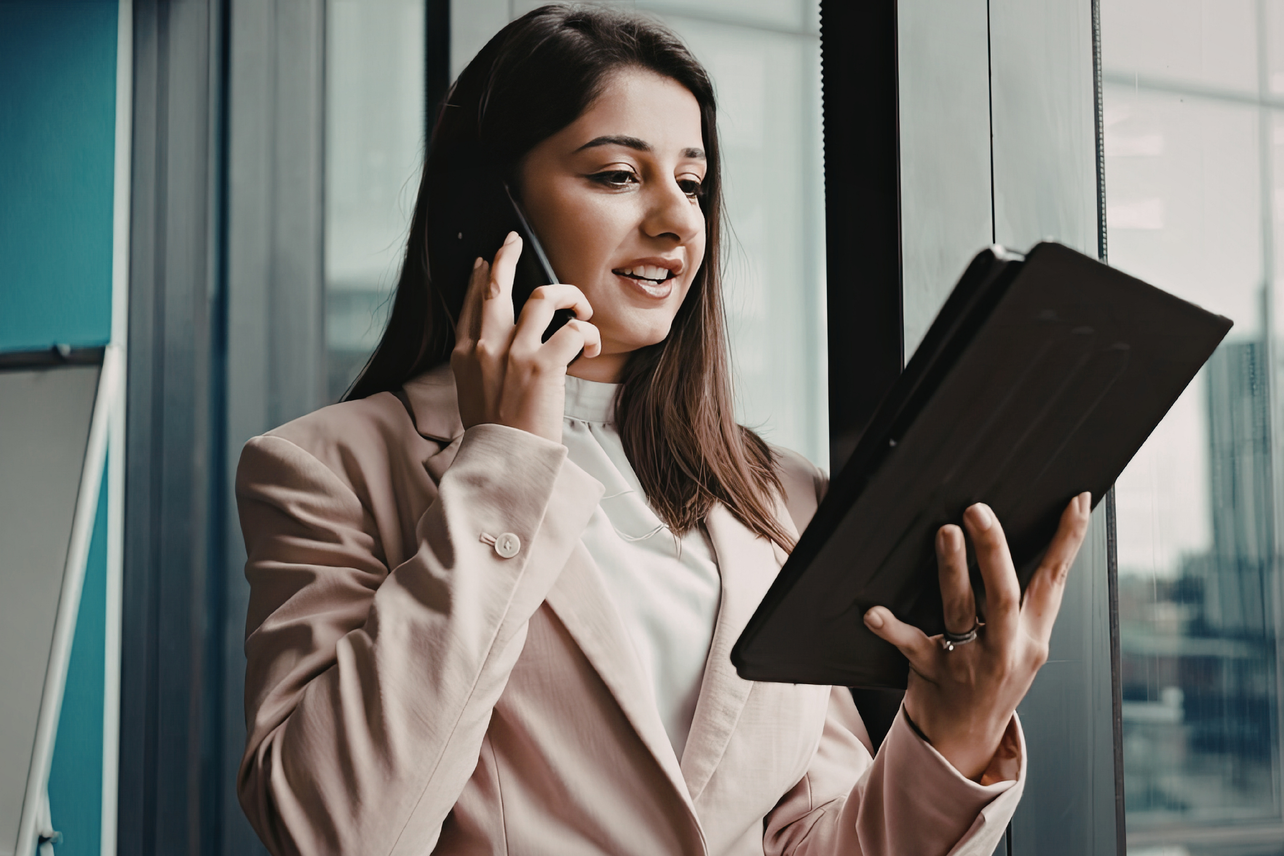 What is VoIP? Business woman wearing a pink blazer and on the cell phone with her right hand, is looking down at a handheld computer with her left hand.