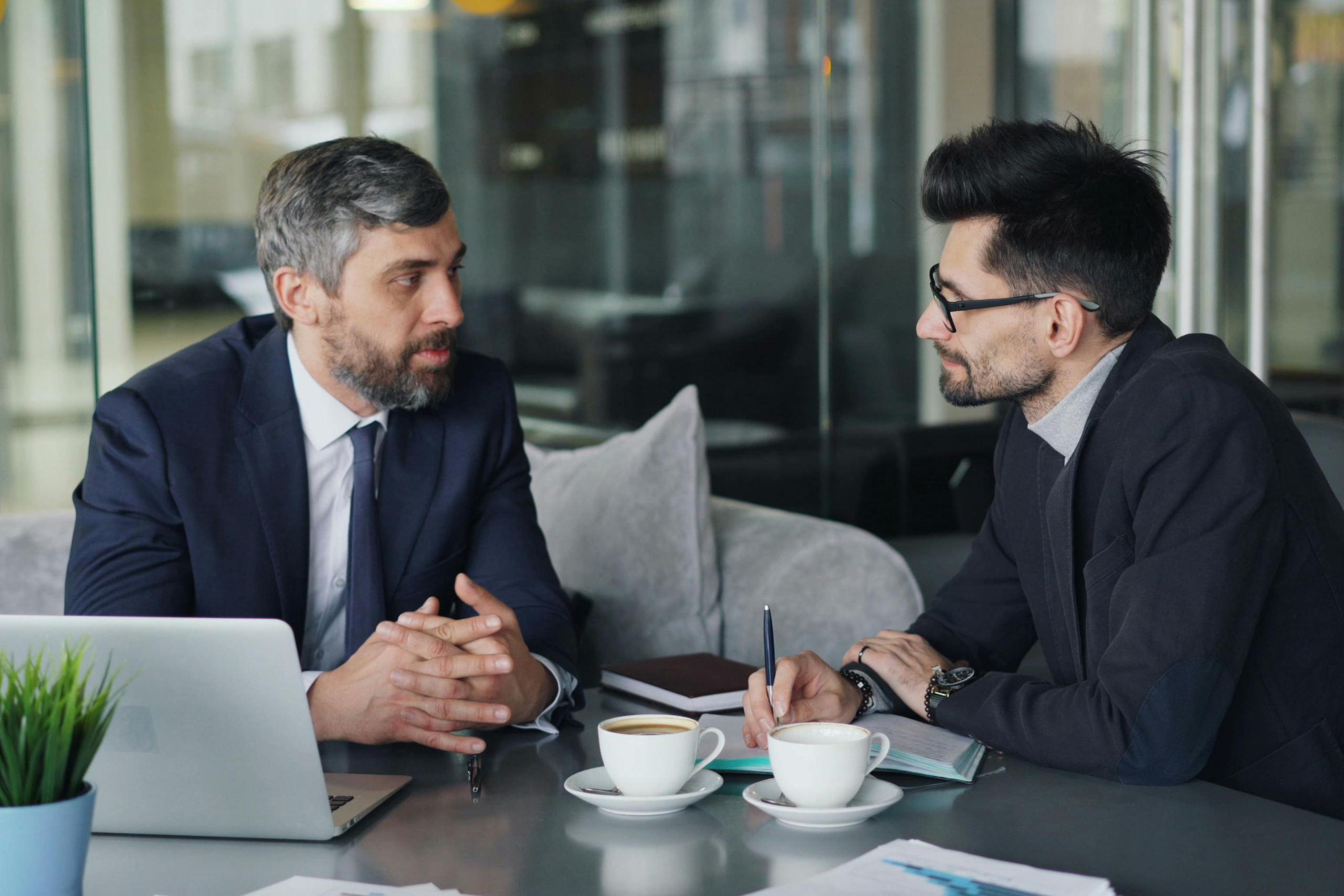 How to talk to your clients switching to voip. This image shows two men in suits sitting down with two coffees in front of them talking to each other. They are presumably having a conversation about switching their VoIP provider.